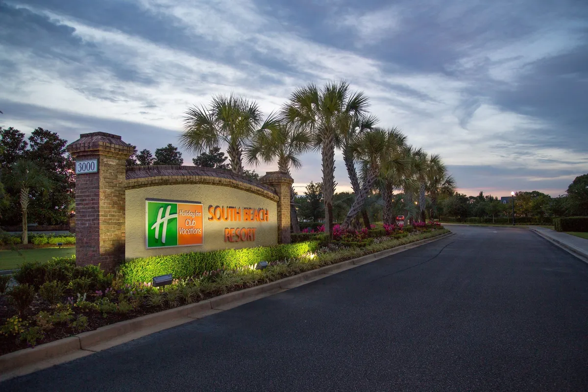 The welcoming entrance to South Beach Resort is lined with palm trees and lush landscaping, featuring the resort sign and inviting pathways. The scene suggests arrival, evening walks, and the overall hospitable atmosphere of the resort grounds.