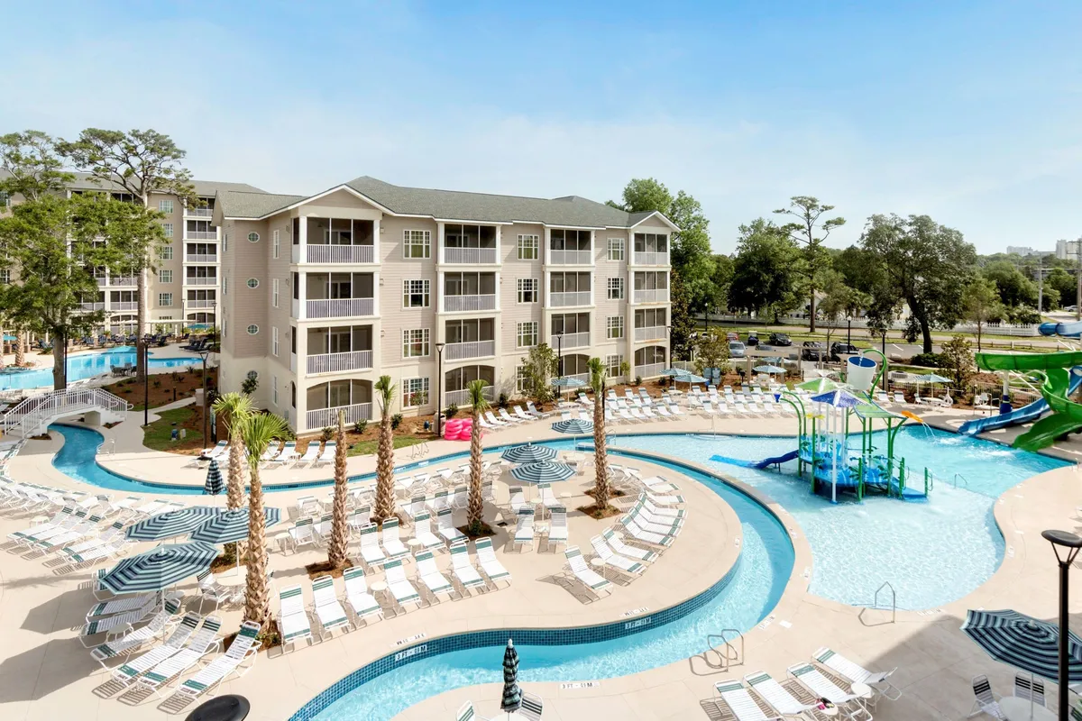 A vibrant outdoor pool scene with families and children enjoying the water, lush palm trees providing shade, giant water slides in the background, and plenty of deck chairs for lounging. The joyful atmosphere is underscored by sunset lighting and visible poolside gatherings.