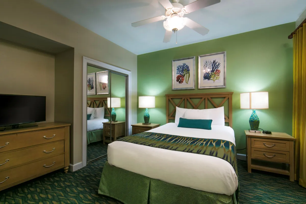 This image features a well-appointed bedroom with a comfortable bed dressed in fresh linens and decorative throw pillows. The space is brightened by natural sunlight streaming through the window, and accented with a green feature wall, vibrant artwork, and turquoise-based lamps. The atmosphere is both inviting and vibrant, suggesting a restful setting for guests.