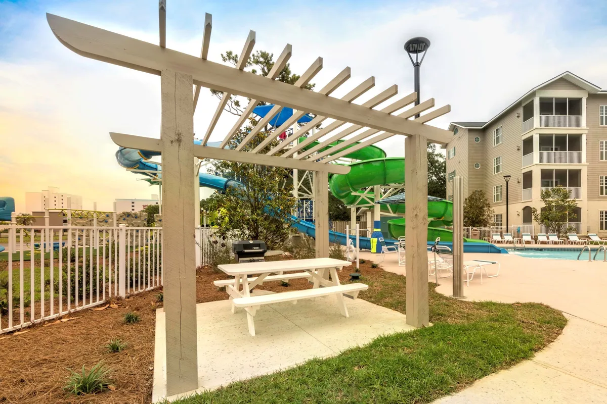 A sunny outdoor scene featuring a lively swimming pool, a water slide, a shaded pergola, and a picnic table. The atmosphere is cheerful, with spaces designed for swimming, sliding, outdoor dining, and socializing—ideal for families or friends enjoying time together.